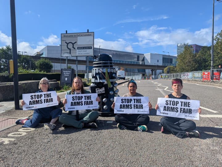 Dalek and Green party Assembly members/councillors holding signs "Stop the Arms Fair."